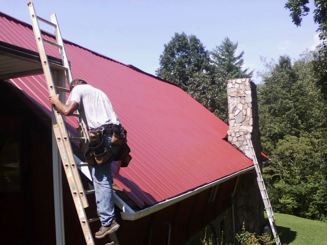 Roofer on a ladder beside a red metal roof and stone chimney — Soffit & Fascia Repair by Roofing Brothers in Cedar Run